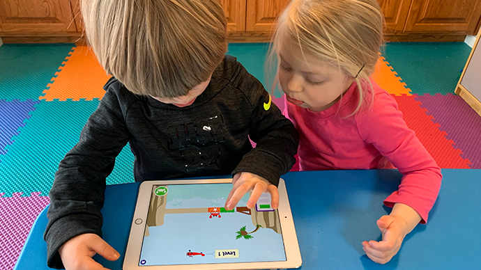 Two children are sitting at a blue table and playing an iPad displaying the Coconut Canyon app, which has a bridge spanning two cliffs and a crab on it.