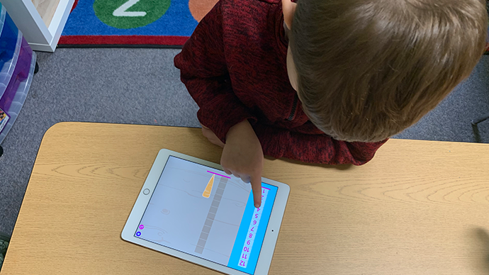 A child is sitting at a laminated wood table and playing an iPad displaying the Farmers Market app, which has a carrot and a measuring ruler on it.