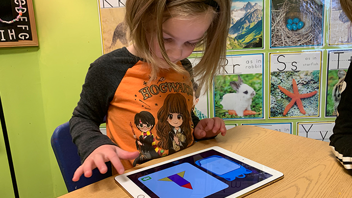 A child is sitting at a laminated wood table and playing an iPad displaying the Shadow Cave app, which has a rocket-shaped puzzle on it made of smaller pieces.