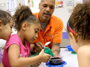 Teacher helping student water a plant while two students watch.