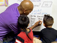 Teacher writing on white board while two students watch.