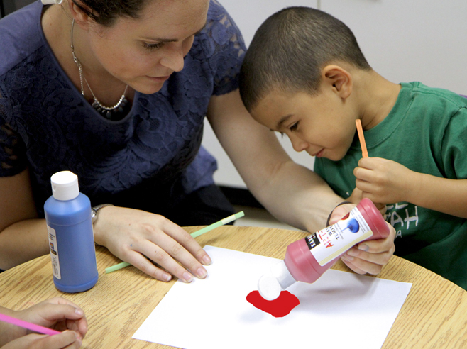 A teacher helps a student squeeze some red paint onto a piece of paper.