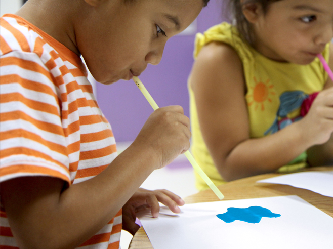 A student blows through a straw to move some blue paint on a piece of paper.