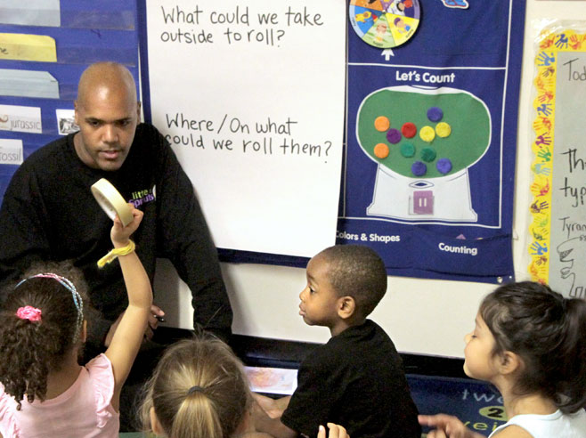 A teacher talking with students seated on the floor. One student is holding up a roll of masking tape.