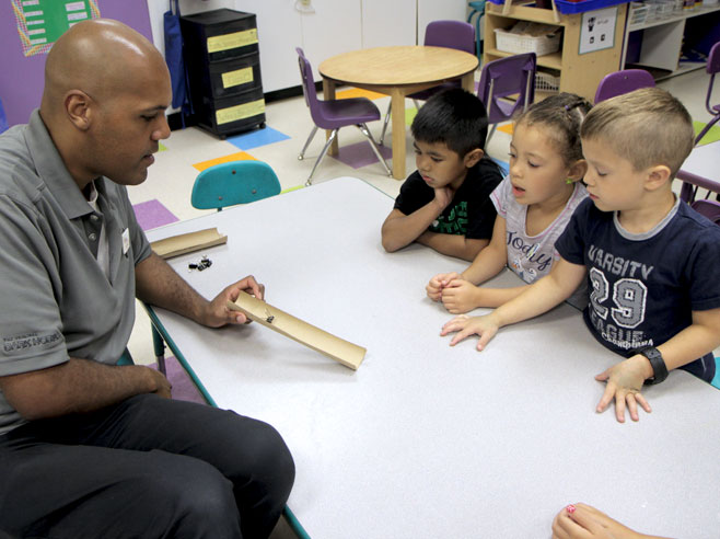 Three students watch teacher slide a binder clip down a ramp made from half a cardboard tube.
