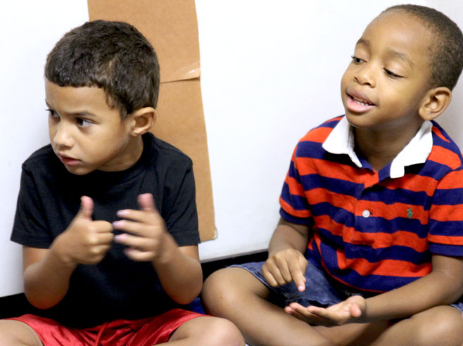 Two students, seated on the floor, making different hand motions. One is clearly talking.