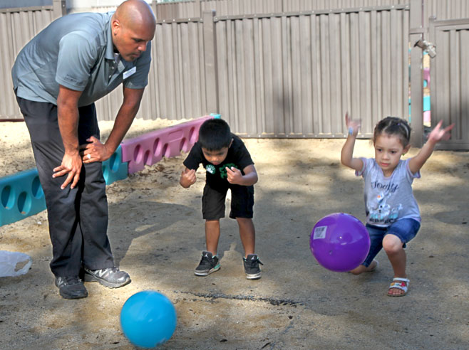 A teacher bends down to watch two students roll large plastic balls across a sandy playground.