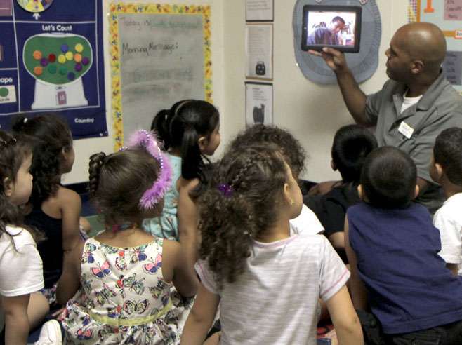 A teacher holds up an iPad to show a class of students a video.