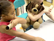 Two students sit at table holding cardboard tubes at the top of their arms, getting ready to use their arms as ramps for the tubes to roll down.