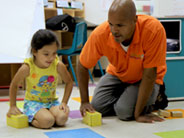 A teacher and a student kneel on the floor. They both are about to push yellow cardboard blocks across the floor.