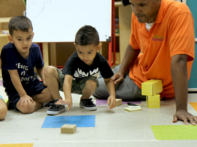 A student pushes a block across the floor, as a teacher and another student look on.