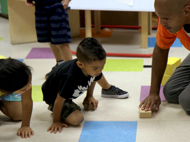 A teacher is about to push a block across the floor, as two students look on.