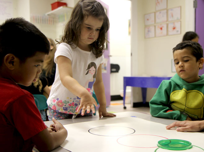 A student stands at a table having just pushed a plastic lid across a poster board with circles drawn on it.
