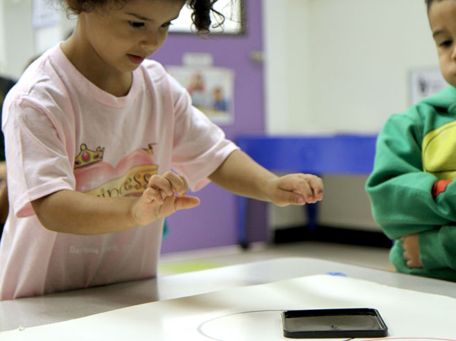 A student stands at a table having just pushed a plastic lid across a poster board with circles drawn on it.