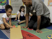 A student and teacher kneel over a gently sloping cardboard ramp. The student holds a pencil in front of two toy cars, to keep them from going down the ramp.