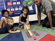 Students and a teacher look on, as a girl rolls a giant marble down a wooden ramp made from cove molding.
