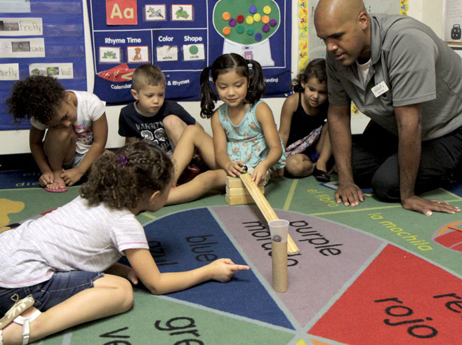 Students and a teacher watch, as a girl rolls a giant marble down a wooden ramp made from cove molding. Another student points to an obstacle that has been set up, at the bottom of the ramp, made from a paper tube with a plastic cup sitting on top of it.