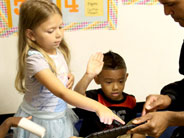 Student points at iPad held by teacher. A boy in the background raises his hand.
