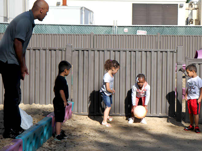 Students and teacher watch as one student prepares to roll a big orange ball.