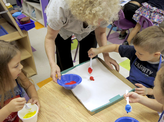 A teacher helps a student spoon a giant marble covered in red paint onto a piece of paper that sits in a tray.