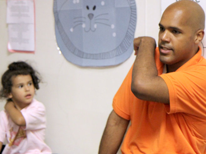 A teacher demonstrates the finger play, 'The Slide', holding his fingers at the top of one shoulder.
