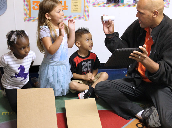 Three students sit behind two differently sloping cardboard ramps. A teacher is showing them a cardboard tube and an iPad.