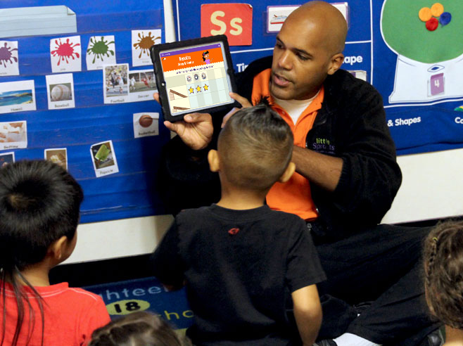 A teacher holds an iPad up and points to the screen. Students look on.