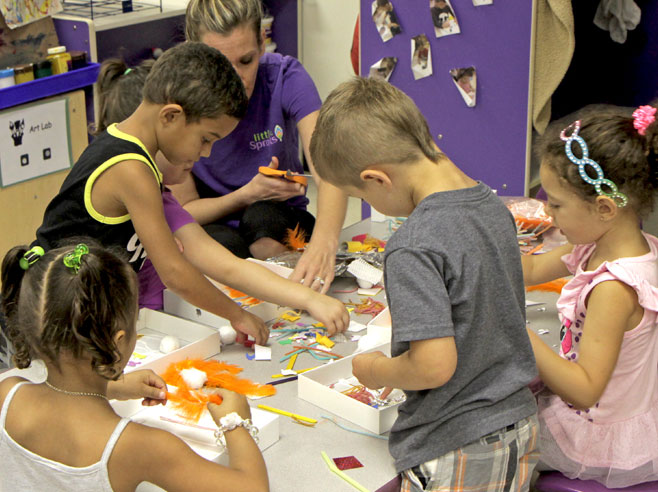 Students and a teacher around a table, work to make collages of differently textured materials in cardboard box lids.