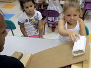 A cardboard ramp supported with wooden blocks is set up on a table. A student slides a block, covered in textured material, down the ramp.
