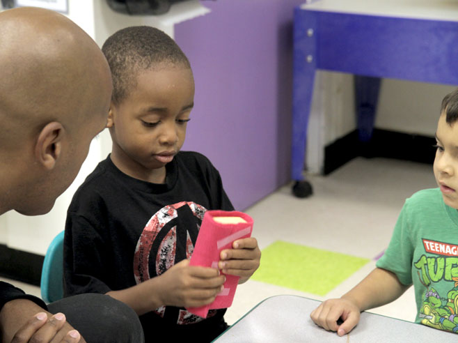 A student examines a block covered in felt. A teacher and another student look on.