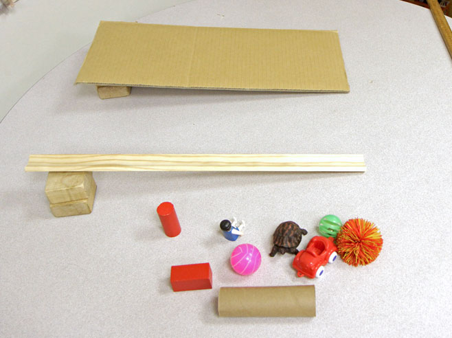 2 ramps sit on a table. One is made from a cove molding and the other is a piece of cardboard. Various toys lie next to the ramps.