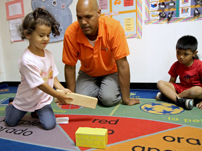 A teacher and student look on, as a girl prepares to use a wooden block to push a cardboard block across the rug.