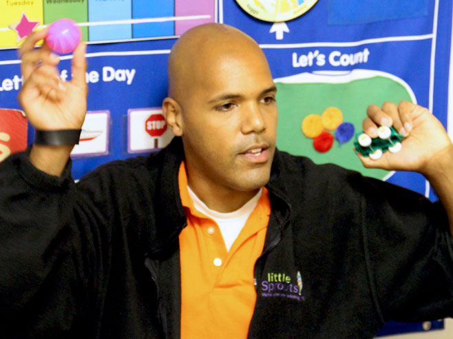 A teacher holds up a pink plastic ball in one hand, and a toy car in the other.