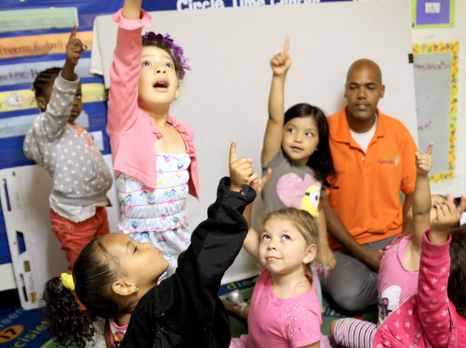 Students, some seated, some standing, point towards the ceiling. A teacher looks on.