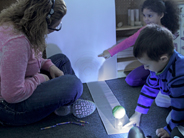 A teacher points to a shadow, created by a tennis ball on top of a cup, which stands on a strip of paper on the floor.