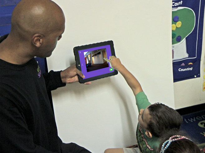 Teacher holds up iPad showing Cool Shadows app screenshot. A student points at the screen.