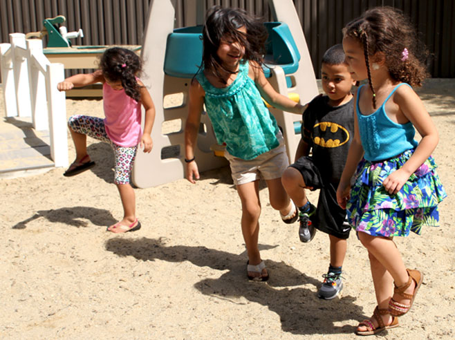 Four students are dancing in a sandy playground. It is sunny, and their shadows are visible.