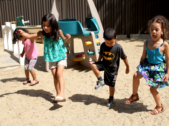 Four students are dancing in a sandy playground. It is sunny, and their shadows are visible.