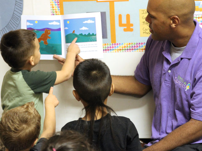 A teacher, seated on the floor, holds up the book 'Moonbear's Shadow'. A student points at an illustration in the book.