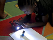 A student crouches on the floor to shine a flashlight, from above, on a small toy that is placed on a piece of white paper.