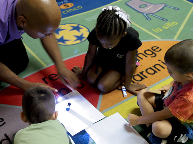 A teacher shines a flashlight on a small toy that is placed on a piece of white paper on the floor. Three students sit on the floor watching.