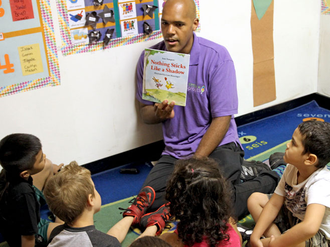 Sitting on the floor, a teacher holds up the book, 'Nothing Sticks Like a Shadow', as a group of students look on.