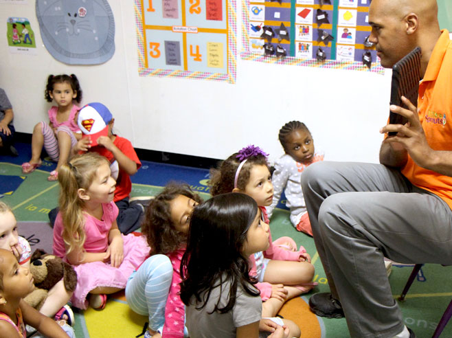 Side view of a teacher sitting on a chair in front of students, who are seated on the floor. He is showing something on an iPad.