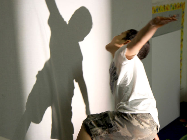 A student dances, and watches his shadow move, on a white wall in front of him.