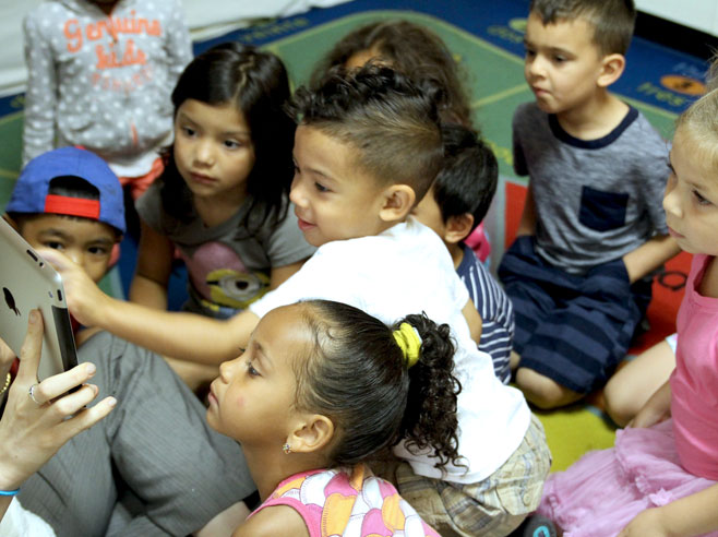 A group of students sit on the floor. One of them points at an iPad being held up in the lower corner of the screen.