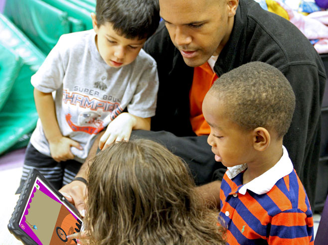 Three students are gathered around a teacher, who is seated on the floor. The teacher holds an iPad showing a Shadow Play app game screenshot. One of the students points at the screen.