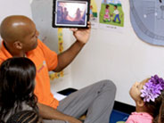 A teacher, seated on the floor, holds up and looks at an iPad, on which a video plays. Students are seated with their backs to the camera, watching the video.