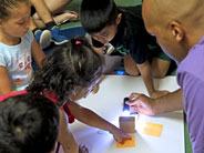 Students gather around a white poster-board on the floor, on top of which is a wooden block with sticky notes placed around it. A teacher points a flashlight at the block as a student points at the shadow created.