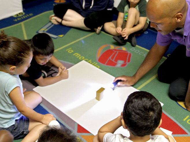 A piece of white poster-board lies on the floor and students are gathered around it. A wooden block sits on the poster-board, and a teacher kneels and shines a flashlight at the block, creating a shadow.