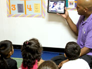 A teacher holds an iPad up in front of a class of students.